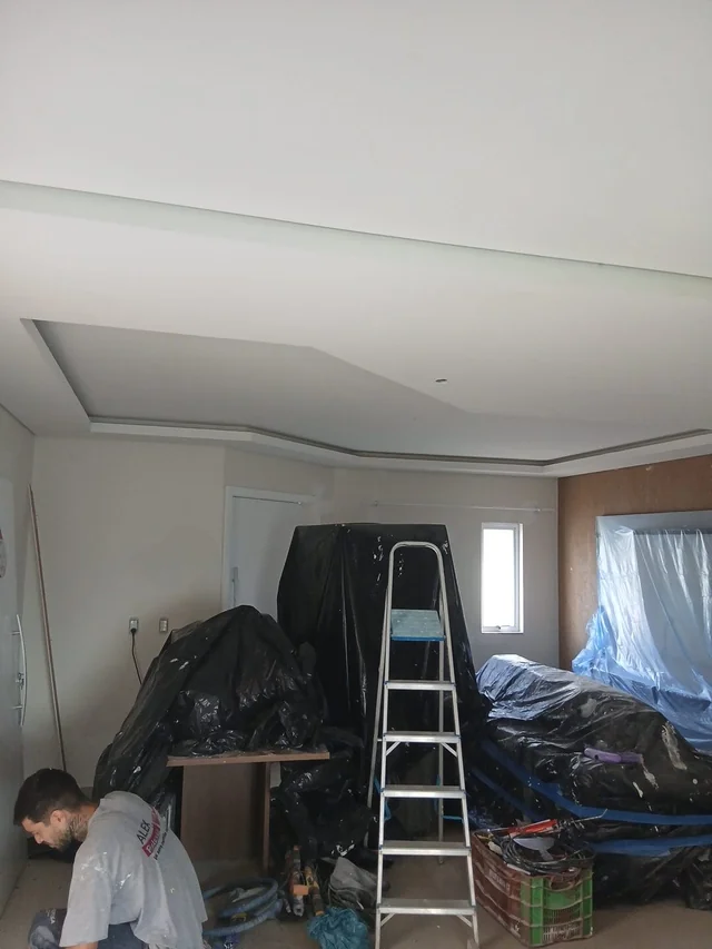 Ambiente em obra com teto rebaixado, ferramentas e móveis protegidos.